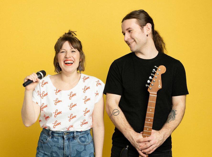 Hymyilevä nainen pitää mikrofonia ja mies kitaraa.